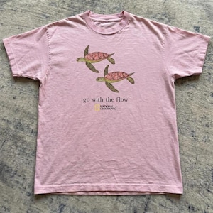 Pu&ograve; includere: Una t-shirt a maniche corte di colore rosa chiaro con una grafica raffigurante due tartarughe marine che nuotano. Sotto le tartarughe, il testo recita "go with the flow" seguito dal logo National Geographic con un rettangolo giallo e testo nero.