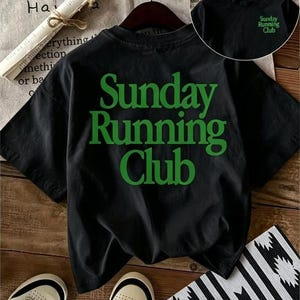 May include: A black short-sleeved t-shirt featuring the text "Sunday Running Club" in large green letters on the back. A smaller version of the same green text is printed on the upper left chest area of the shirt, which is displayed on a hanger.