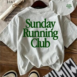 May include: A light gray short-sleeve t-shirt featuring the dark green text "Sunday Running Club" printed in a large font on the back and a smaller version of the same text on the front chest area, displayed on a hanger with a crew neckline.