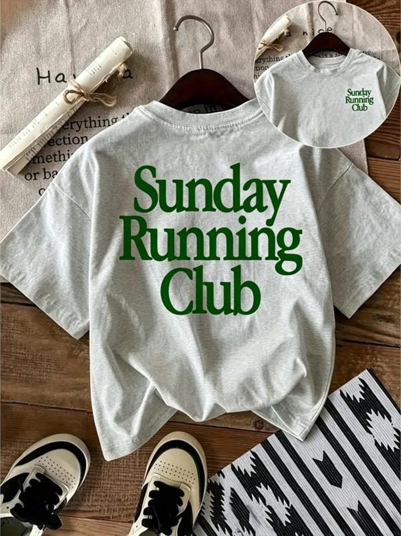 May include: A light gray short-sleeve t-shirt featuring the dark green text "Sunday Running Club" printed in a large font on the back and a smaller version of the same text on the front chest area, displayed on a hanger with a crew neckline.