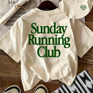 May include: An off-white short-sleeve t-shirt featuring the dark green text "Sunday Running Club" printed in a large serif font on the back. A smaller version of the same green text is printed on the upper left chest area of the shirt.