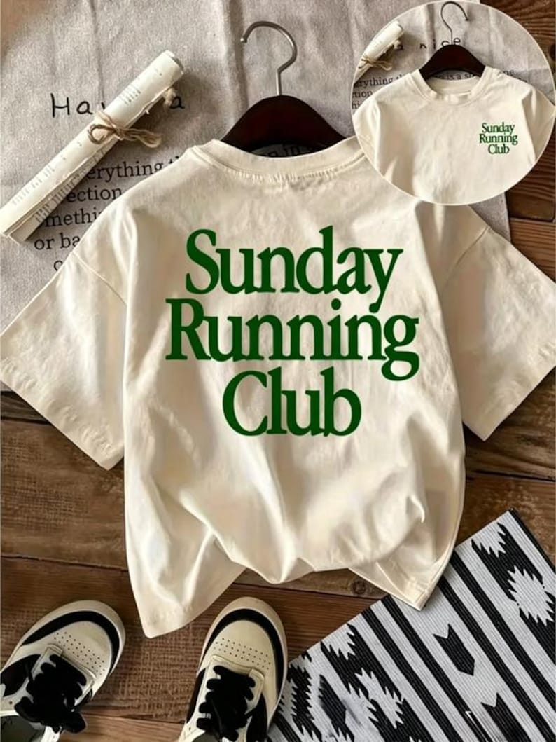 May include: An off-white short-sleeve t-shirt featuring the dark green text "Sunday Running Club" printed in a large serif font on the back. A smaller version of the same green text is printed on the upper left chest area of the shirt.