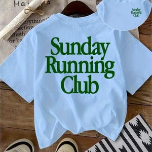 May include: A light blue short-sleeve t-shirt featuring the dark green text "Sunday Running Club" printed in a large font on the back. A smaller version of the same green text is printed on the upper left chest area of the shirt.
