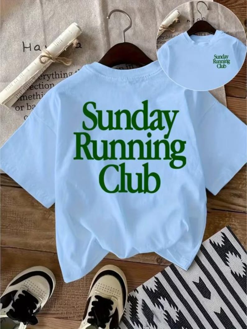 May include: A light blue short-sleeve t-shirt featuring the dark green text "Sunday Running Club" printed in a large font on the back. A smaller version of the same green text is printed on the upper left chest area of the shirt.