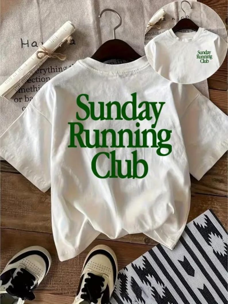 May include: A white short-sleeve t-shirt featuring the text "Sunday Running Club" in large dark green letters on the back. A smaller version of the same green text is printed on the front chest area of the shirt, which is displayed on a hanger.