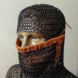 May include: A black chainmail coif covering the head and neck, featuring a tan leather trim that frames the nose and mouth area. The metal rings are linked in a dense pattern, providing full coverage while leaving the eyes visible.