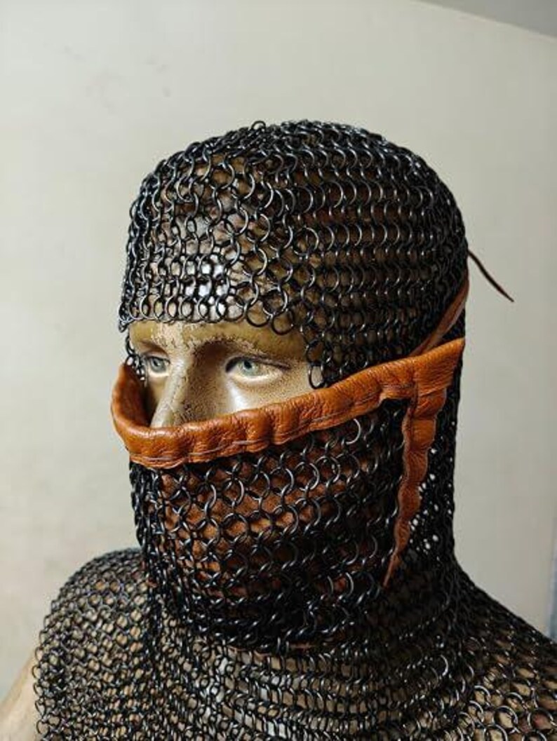 May include: A black chainmail coif covering the head and neck, featuring a tan leather trim that frames the nose and mouth area. The metal rings are linked in a dense pattern, providing full coverage while leaving the eyes visible.
