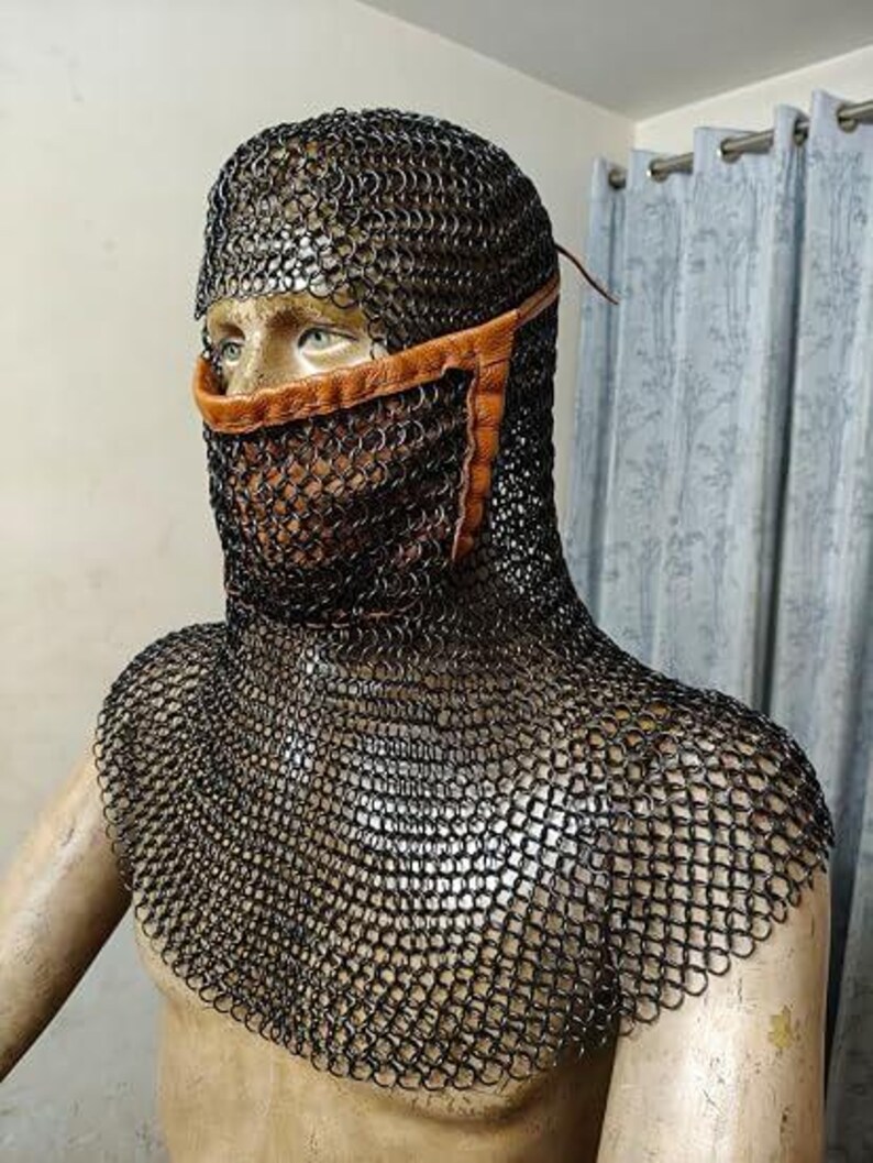 May include: A black chainmail coif featuring a tan leather face mask integrated into the metal rings. The hood covers the head and shoulders, constructed from interlocking dark metal links, with a leather strap securing the nose and mouth area.