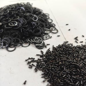 May include: A close-up shot of a pile of small, black metal washers and a pile of small, black metal screws. The washers are circular with a hole in the center and a series of notches around the edge. The screws are cylindrical with a slotted head.