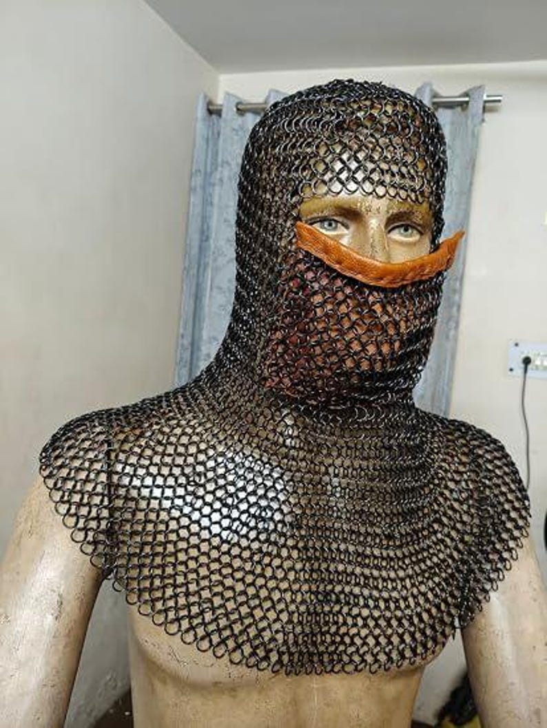 May include: A black chainmail coif featuring interlocking metal rings that cover the head, neck, and shoulders. The headpiece includes a horizontal tan leather strap across the nose area, leaving the eyes exposed while covering the lower face.