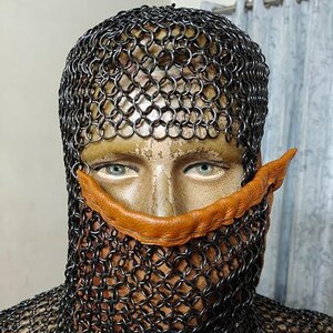 May include: A chainmail coif covering the head and neck, featuring a dark metallic finish with interlocking circular links. A tan, textured leather strip is positioned horizontally across the nose bridge area, resting against the chainmail structure.