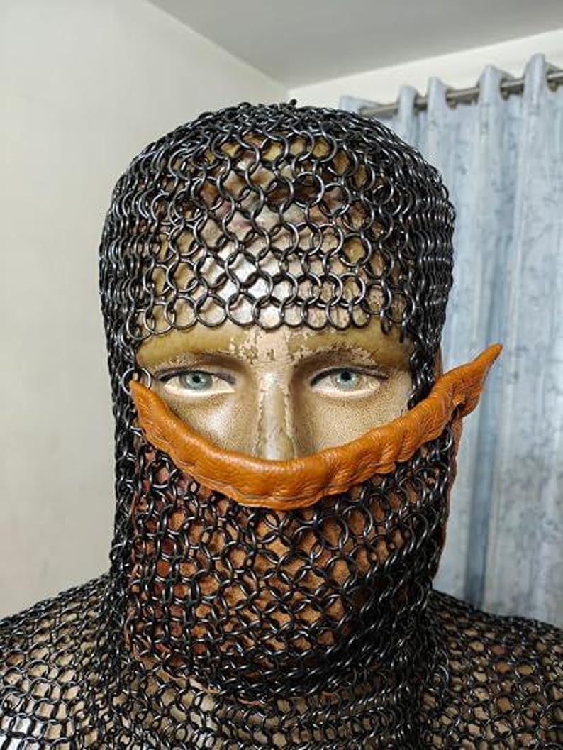 May include: A chainmail coif covering the head and neck, featuring a dark metallic finish with interlocking circular links. A tan, textured leather strip is positioned horizontally across the nose bridge area, resting against the chainmail structure.