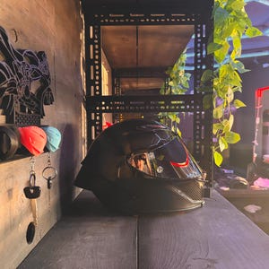 Pode incluir: Um capacete de motocicleta preto com viseira fum&ecirc; est&aacute; sobre uma superf&iacute;cie escura. Um porta-chaves de parede com tampas de chave vermelhas e azuis e chaves est&aacute; pendurado &agrave; esquerda. O fundo inclui prateleiras e vegeta&ccedil;&atilde;o.