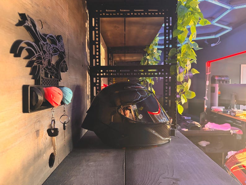 Pode incluir: Um capacete de motocicleta preto com viseira fum&ecirc; est&aacute; sobre uma superf&iacute;cie escura. Um porta-chaves de parede com tampas de chave vermelhas e azuis e chaves est&aacute; pendurado &agrave; esquerda. O fundo inclui prateleiras e vegeta&ccedil;&atilde;o.