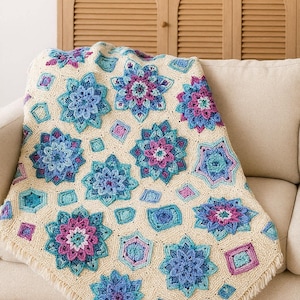 Può includere: Una coperta all'uncinetto color crema con un motivo floreale nei toni del blu, turchese e viola. La coperta è drappeggiata su un divano beige, mettendo in risalto il suo design intricato e i bordi sfrangiati. I motivi floreali sono disposti in un motivo ripetuto.