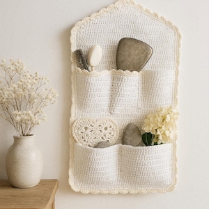 Könnte beinhalten: Ein weißer Wandorganizer aus Häkelarbeit mit mehreren Taschen, der eine Bürste, Seife, Steine und Blumen enthält. Der Organizer hängt an einer weißen Wand. Das Design umfasst einen gewellten Rand und eine Aufhängeschlaufe.