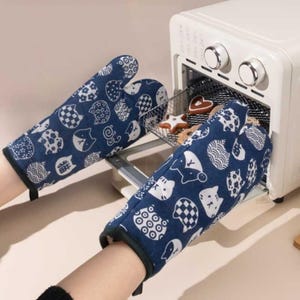 May include: Navy blue oven mitts with a white cat and geometric pattern. The mitts are being used to remove a tray of baked goods from a white oven. The mitts have a black trim and a loop for hanging.