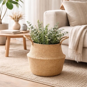 May include: A woven, light brown basket with two handles, filled with green plants. The basket sits on a beige rug in a living room setting. A beige sofa and wooden table are in the background.