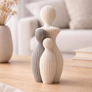 Peut inclure: Ensemble de trois sculptures abstraites, évoquant une famille, en nuances de gris et blanc cassé. Les sculptures présentent des détails verticaux côtelés et reposent sur une surface en bois clair. Un élément de décoration intérieure.