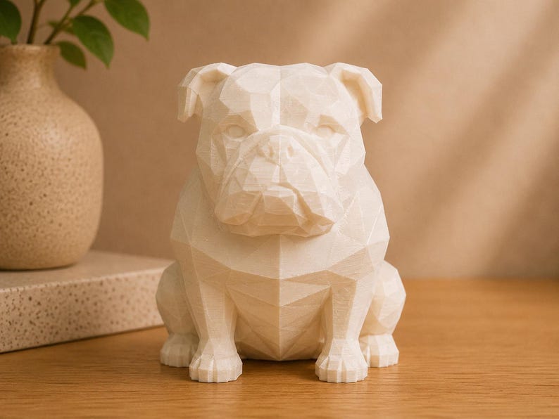 May include: A cream-colored, low-poly geometric bulldog figurine featuring a faceted, angular surface design. The sculpture is rendered in a solid, matte material and depicts the dog in a seated, forward-facing position with distinct ears.