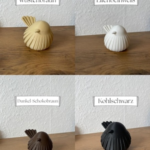 May include: Four small bird figurines in different colors. The top left is W&uuml;stenbraun, the top right is Elfenbeinwei&szlig;, the bottom left is Dunkel-Schokobraun, and the bottom right is Kohlschwarz. Each bird has a rounded body with vertical ridges.