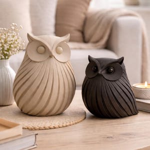 以下が含まれることがあります： ライトベージュ仕上げとダークチャコール仕上げの2体のフクロウの置物。どちらも滑らかで丸みを帯びた体に垂直の彫り線が施されており、顔には目立つ円形の目のディテールが特徴的です。