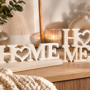 以下が含まれることがあります： 「HOME」という文字をかたどった2つの白い木製装飾品。1つは文字が横並びで、「O」の代わりにハート型が配置されています。もう1つは「H」が「M」と「E」の上に配置された縦型のデザインです。