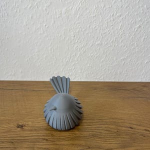 May include: A small, matte gray bird figurine featuring a rounded body with a series of vertical, fan-like ridges along its sides and a prominent, upright, fan-shaped tail. The bird has a smooth, conical head and a small, pointed beak facing forward.