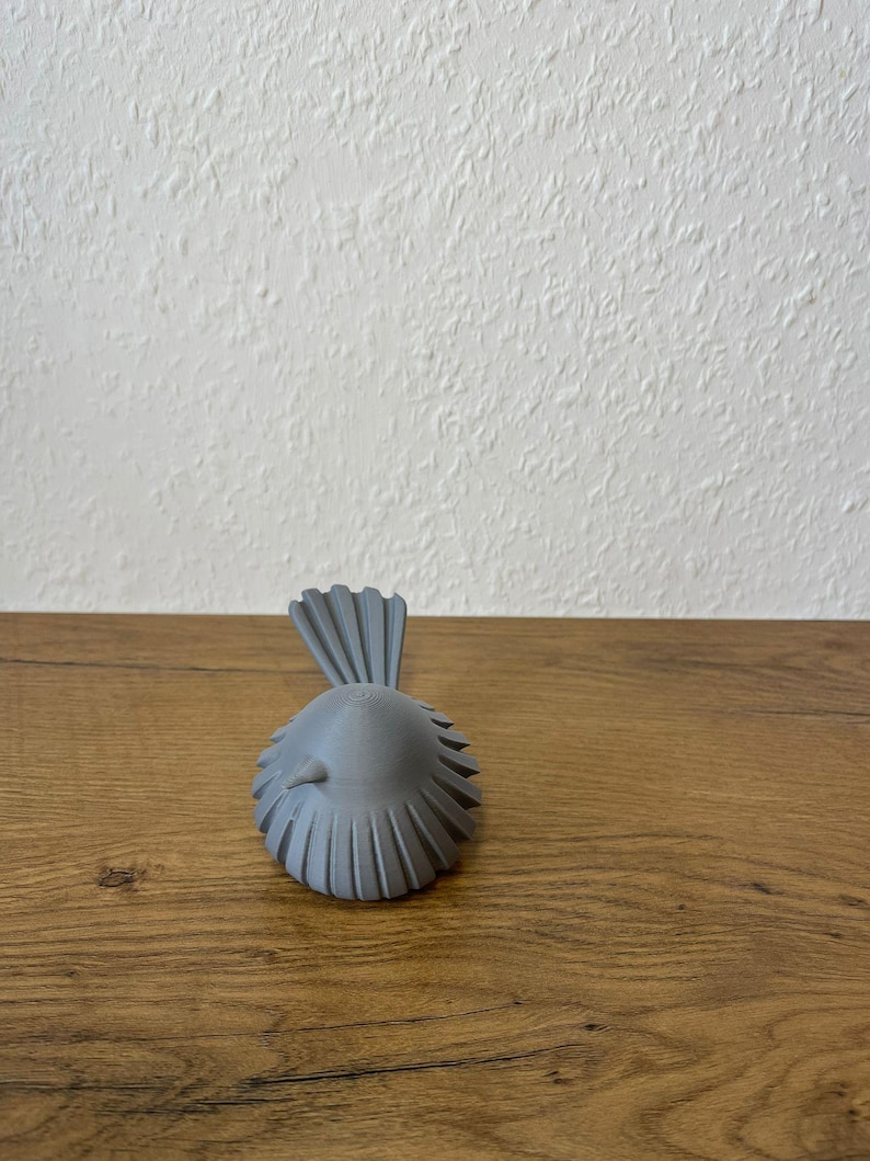 May include: A small, matte gray bird figurine featuring a rounded body with a series of vertical, fan-like ridges along its sides and a prominent, upright, fan-shaped tail. The bird has a smooth, conical head and a small, pointed beak facing forward.