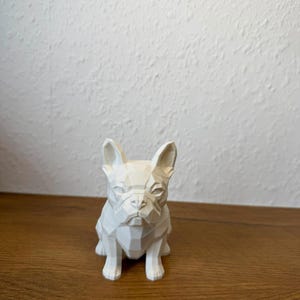 May include: A white, geometric-style figurine of a French Bulldog. The sculpture features sharp, angular facets that define the dog's ears, snout, and body, creating a low-poly appearance. The surface is matte and solid white throughout.