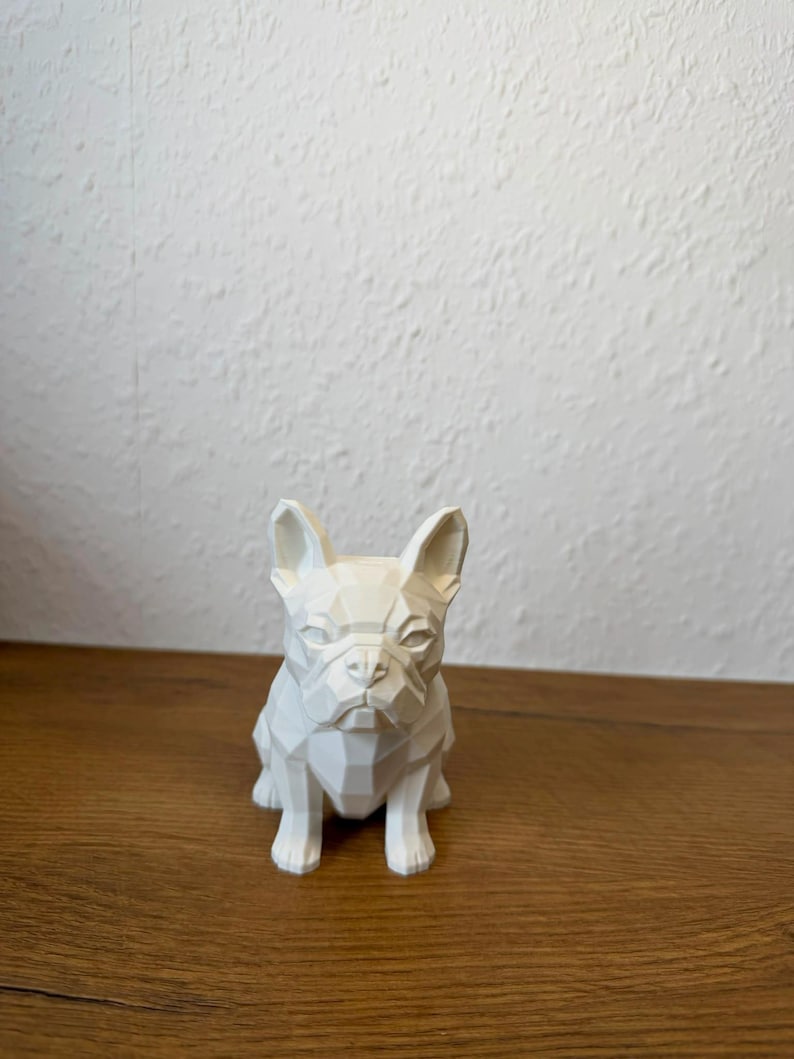 May include: A white, geometric-style figurine of a French Bulldog. The sculpture features sharp, angular facets that define the dog's ears, snout, and body, creating a low-poly appearance. The surface is matte and solid white throughout.