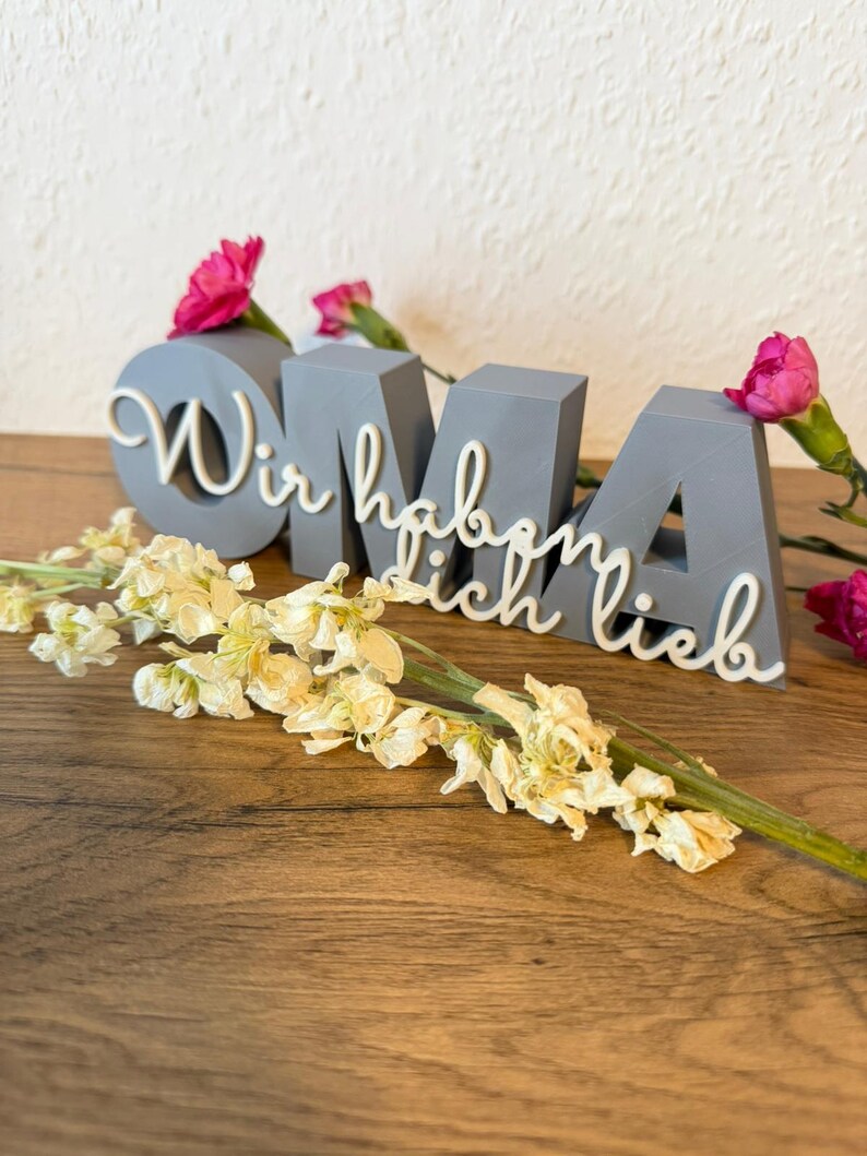 K&ouml;nnte beinhalten: Ein graues, 3D-gedrucktes Schild mit der Aufschrift "OMA" und den Worten "Wir haben dich lieb" in wei&szlig;er Schrift. Das Schild ist mit rosa Nelken und einem Zweig wei&szlig;er Blumen verziert, die auf einer Holzoberfl&auml;che liegen.