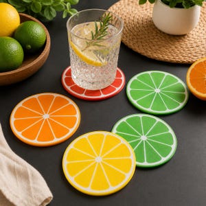 May include: A set of four circular drink coasters featuring vibrant citrus fruit slice designs. The coasters include one orange, one yellow lemon, and two green lime patterns, each with white segment lines on a dark surface.