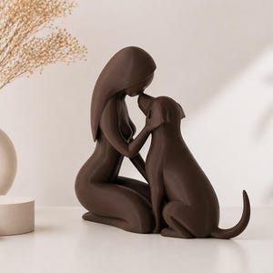 May include: A dark brown sculpture depicting a kneeling figure with long hair touching foreheads with a dog. The figure has a smooth, stylized form, and the dog is shown in a seated position with its tail slightly curved upward, creating a bond.