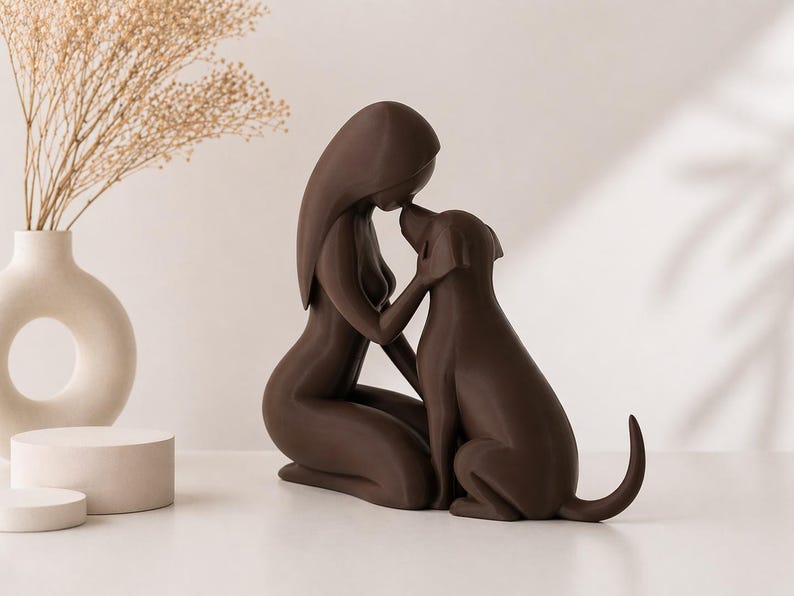 May include: A dark brown sculpture depicting a kneeling figure with long hair touching foreheads with a dog. The figure has a smooth, stylized form, and the dog is shown in a seated position with its tail slightly curved upward, creating a bond.