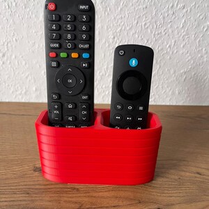 May include: A bright red, oval-shaped remote control holder with horizontal ribbed detailing. The holder contains two black remote controls, one larger with a full numeric keypad and one smaller with a circular navigation pad and microphone icon.