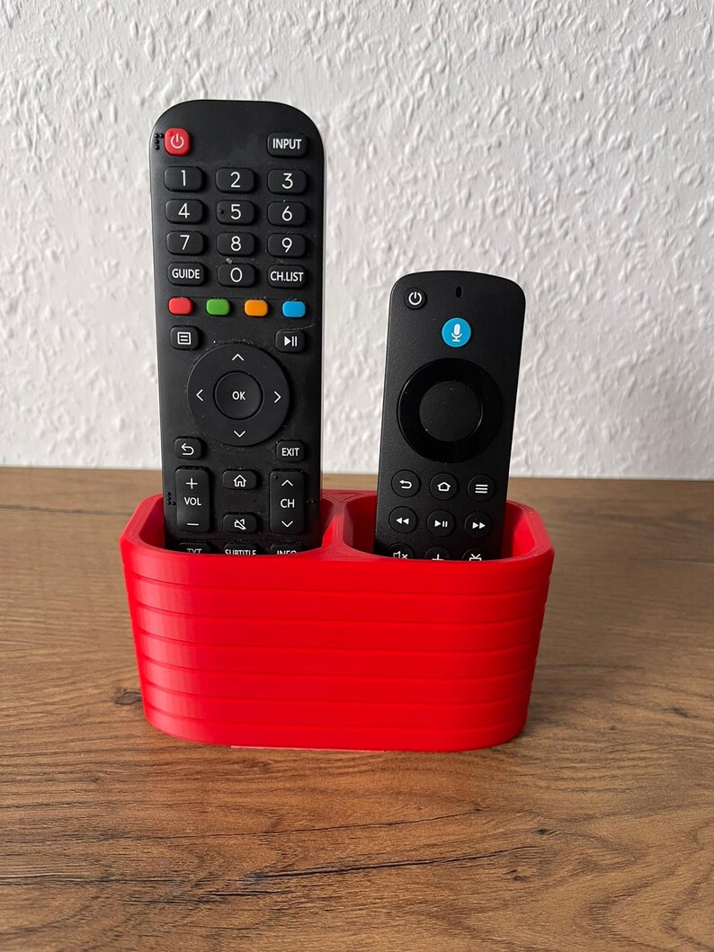 May include: A bright red, oval-shaped remote control holder with horizontal ribbed detailing. The holder contains two black remote controls, one larger with a full numeric keypad and one smaller with a circular navigation pad and microphone icon.