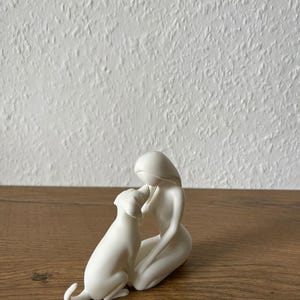 May include: A matte white ceramic figurine depicting a stylized human figure kneeling and interacting with a dog. The smooth, minimalist sculpture features fluid lines and a solid white finish, capturing the form of a person and a canine companion.