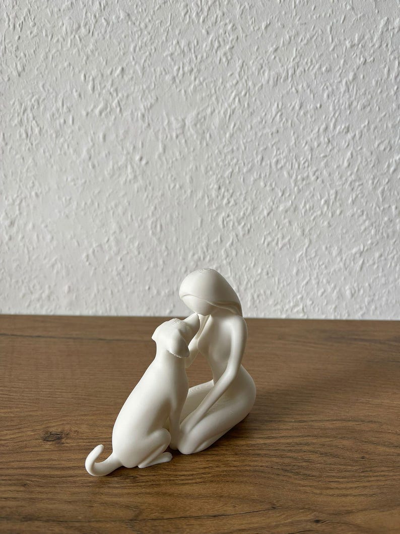 May include: A matte white ceramic figurine depicting a stylized human figure kneeling and interacting with a dog. The smooth, minimalist sculpture features fluid lines and a solid white finish, capturing the form of a person and a canine companion.