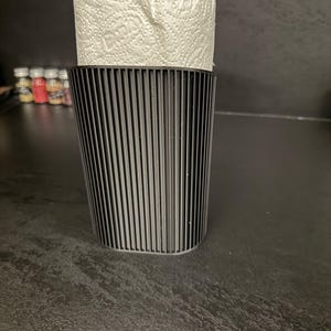 May include: A black, vertically ribbed, cylindrical holder containing a roll of white paper towels with a visible embossed pattern. The holder features a consistent, textured, linear design that wraps around the exterior of the container.