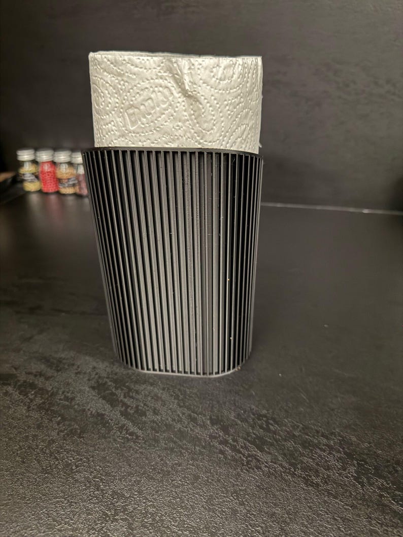 May include: A black, vertically ribbed, cylindrical holder containing a roll of white paper towels with a visible embossed pattern. The holder features a consistent, textured, linear design that wraps around the exterior of the container.