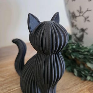 May include: A matte black cat figurine featuring a distinct ribbed or layered texture throughout its body. The cat is depicted in a seated position with its tail curved upward, showcasing vertical lines that define its form and pointed ears.