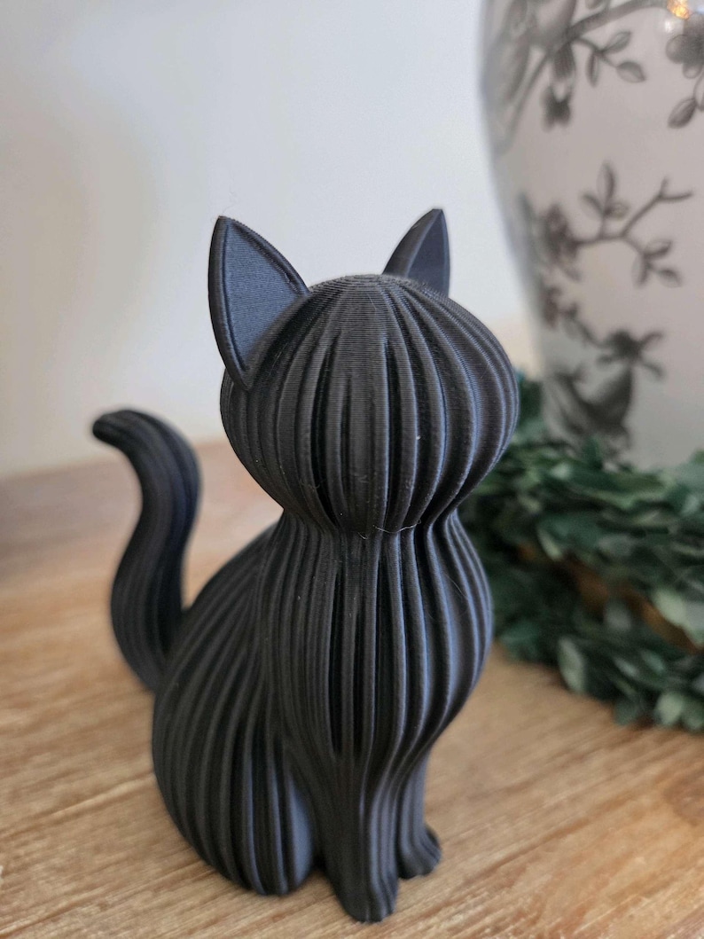 May include: A matte black cat figurine featuring a distinct ribbed or layered texture throughout its body. The cat is depicted in a seated position with its tail curved upward, showcasing vertical lines that define its form and pointed ears.