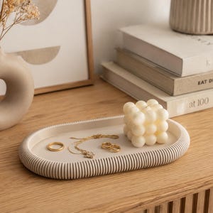 Puede incluir: Una bandeja ovalada beige con borde acanalado contiene joyas de oro y una vela de burbujas blanca. Un jarr&oacute;n beige con flores secas y libros est&aacute;n en el fondo. La bandeja est&aacute; sobre una superficie de madera.
