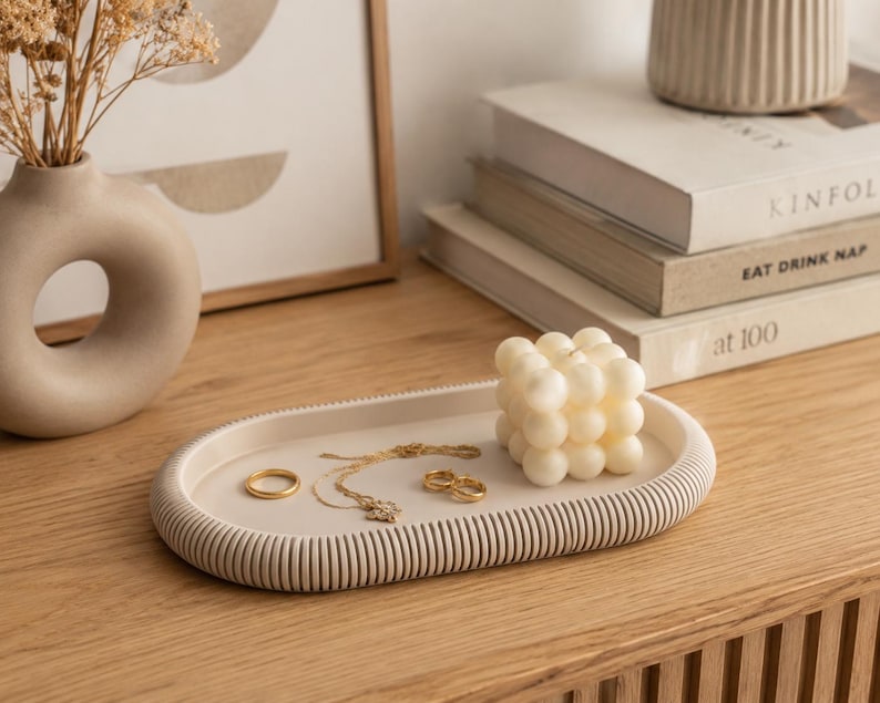 Puede incluir: Una bandeja ovalada beige con borde acanalado contiene joyas de oro y una vela de burbujas blanca. Un jarr&oacute;n beige con flores secas y libros est&aacute;n en el fondo. La bandeja est&aacute; sobre una superficie de madera.