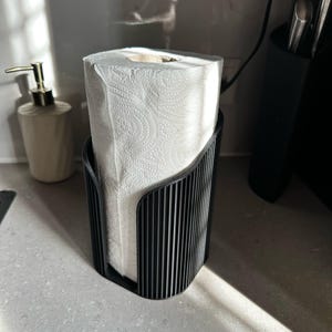 May include: A black, vertically ribbed, cylindrical paper towel holder containing a roll of white paper towels with a textured pattern. The holder features an open-front design that allows for easy access to the paper towels.
