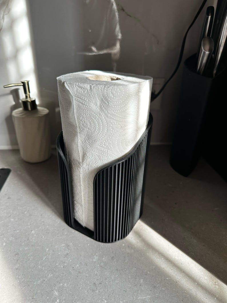 May include: A black, vertically ribbed, cylindrical paper towel holder containing a roll of white paper towels with a textured pattern. The holder features an open-front design that allows for easy access to the paper towels.
