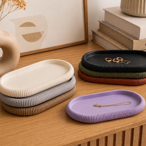 Puede incluir: Bandejas ovaladas en crema, gris, marr&oacute;n, negro, verde y morado. Cada bandeja tiene un borde acanalado. Las bandejas est&aacute;n apiladas sobre una superficie de madera, algunas con joyas. Ideales para guardar objetos peque&ntilde;os.