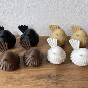 May include: Eight small bird figurines arranged in pairs, featuring two black, two brown, two tan, and two white birds. Each bird has a rounded body with vertical ribbed detailing and a fanned tail, all crafted with a smooth, matte finish.