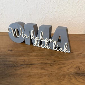 K&ouml;nnte beinhalten: Ein graues, 3D-gedrucktes Schild mit dem Wort "OMA" in Blockbuchstaben, mit dem kursiven Text "Wir haben dich lieb" dar&uuml;ber. Das Schild steht auf einer Holzoberfl&auml;che.